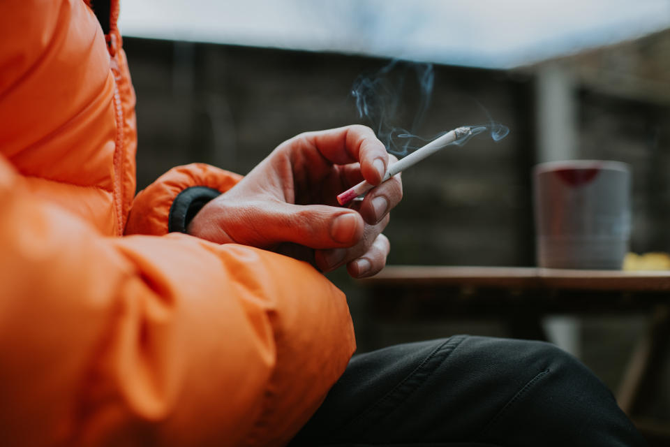 A man sits outside and holds a thin menthol cigarette between his fingers while smoking.  Close-up with focus on the cigarette.  (Photo via Getty Images)