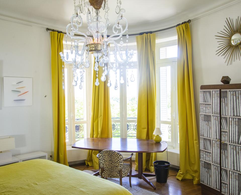 On the ceiling hangs a Spanish chandelier from the 18th century. The table is by Italian architect and designer Franco Albini for Poggi, circa 1950.  The cupboard is from the 1930s covered with Fornasetti wallpaper, and on the wall is an artwork by British artist Marc Cavell. A 1930s chair is covered in velvet, and the sun mirror is from the Paris flea market.