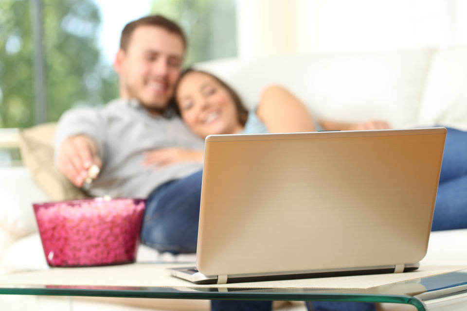 A young couple cuddle on the couch with a bowl of popcorn and a laptop.