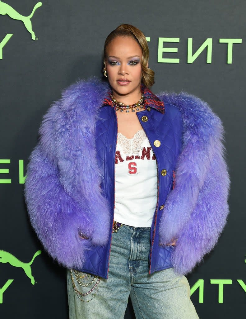 Rihanna in a puffy jacket over a graphic tee, with layered necklaces and jeans, posing for the camera