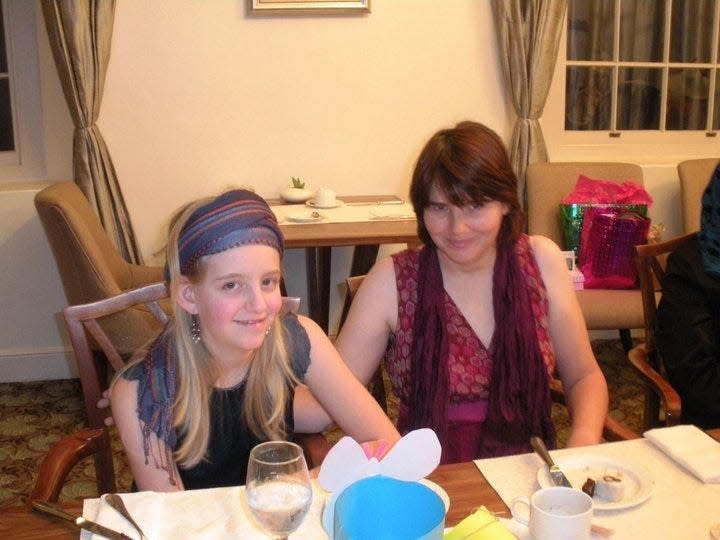 Nicci with her daughter Danny, seated at a restaurant. Danny wears a blue striped scarf wrapped around her head. She has long blonde hair and wears a black tank top. Nicci has chin-length dark hair and wears a magenta top with a graphic pattern. She has her arm on Danny. Both smile at the camera.