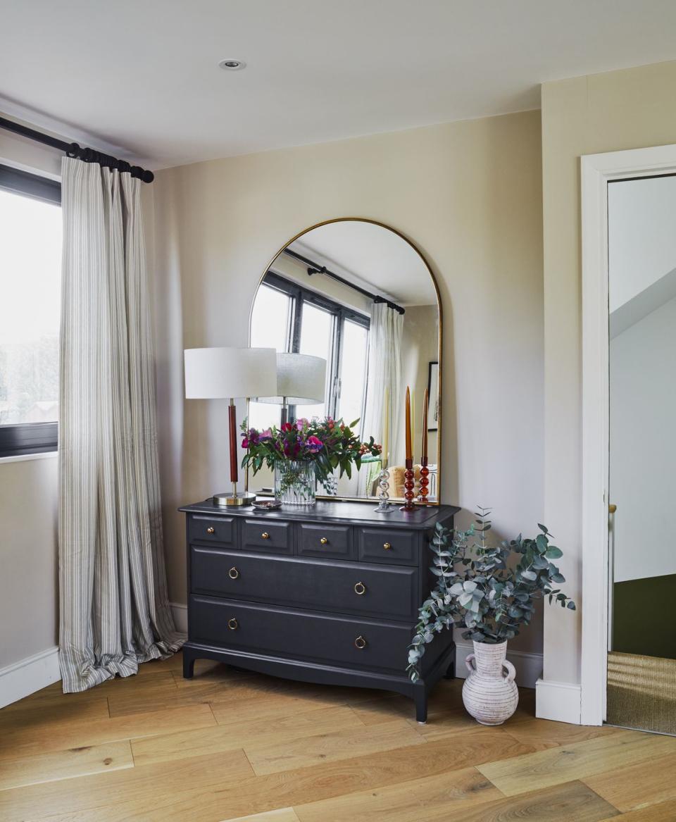 black chest of drawers in corner of bedroom with an arched mirror and lamp on top