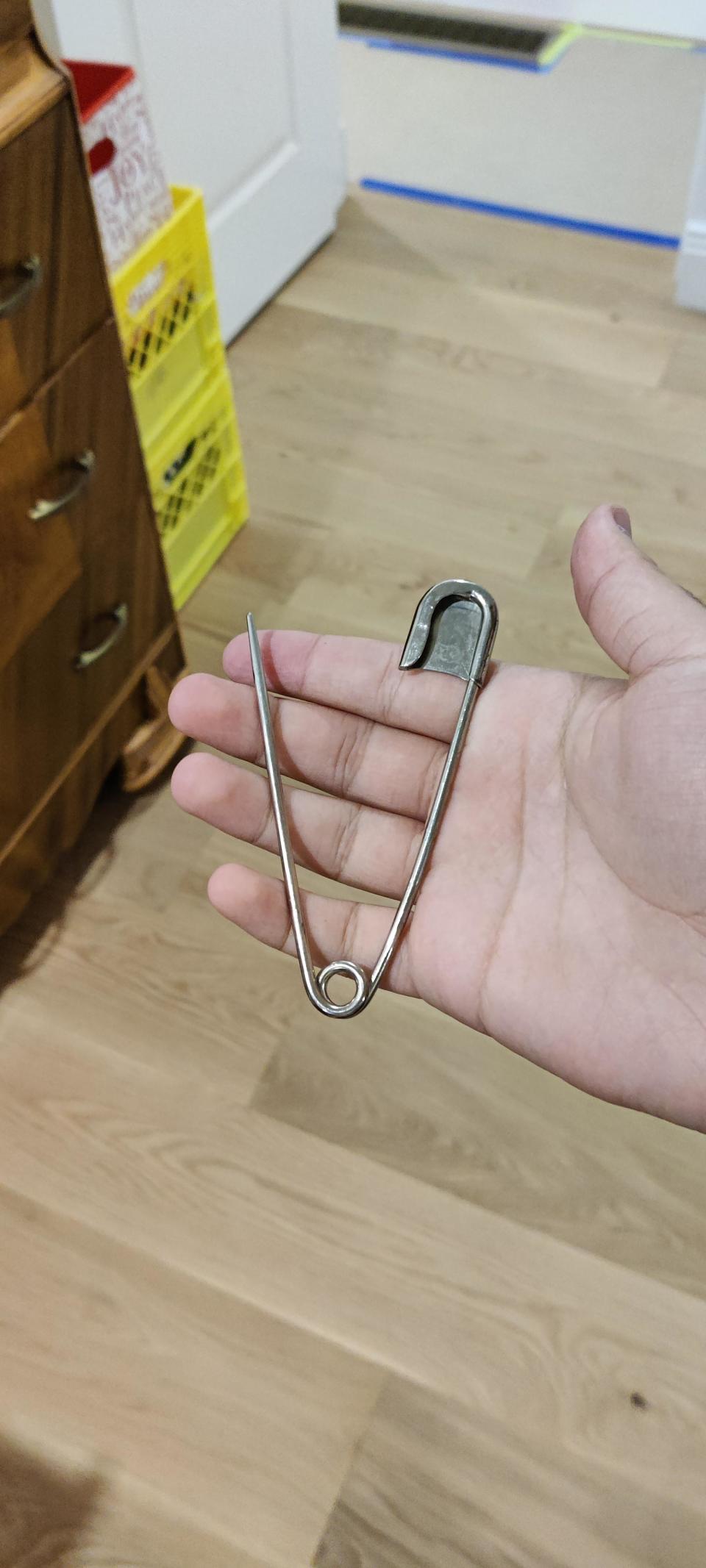 A person's hand holding a large metal safety pin against a home interior background