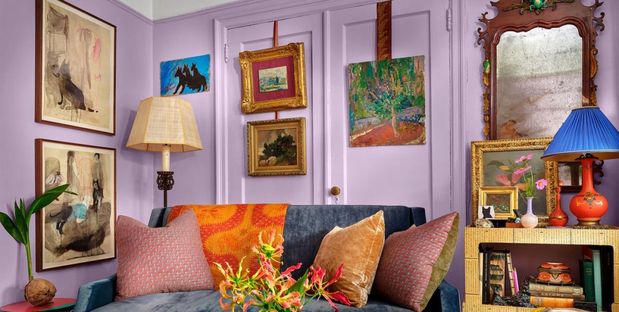 a purple living room with artwork on the walls and a blue velvet covered sofa at center with multiple colored pillows on it and a shelf next to it with books and objects and a large mirror behind it