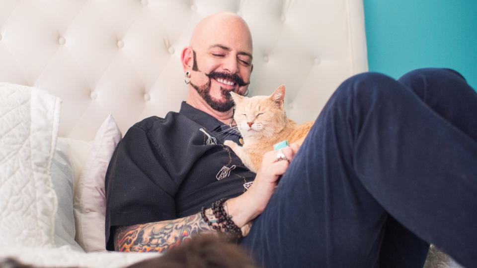 Jackson Galaxy sits on a couch and cuddles an orange tabby cat. (Photo by Andrew Marttila.)