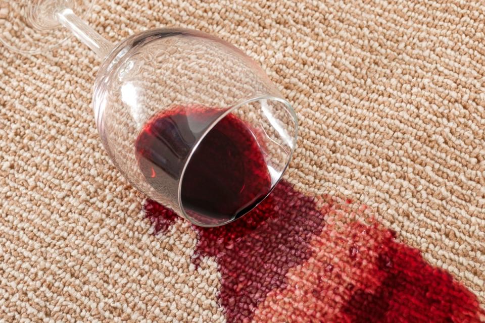 Wine glass sideways on a beige carpet, with red wine spilling out onto the carpet.