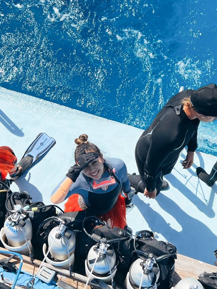 Three people in scuba gear on a dock. A woman in the center wearing a pink tutu guard looks up and smiles. A man in black scuba gear stands next to her