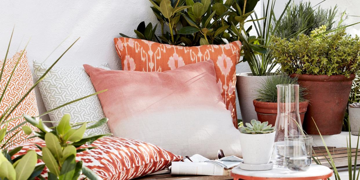 outside seating area, white bench with a wooden top covered in orange patterned cushions and a small table surrounded by pot plants instant oasis transform a shaded garden nook with piles of ikat print cushions in warming hues of burnt ochre and planters filled with lush foliage