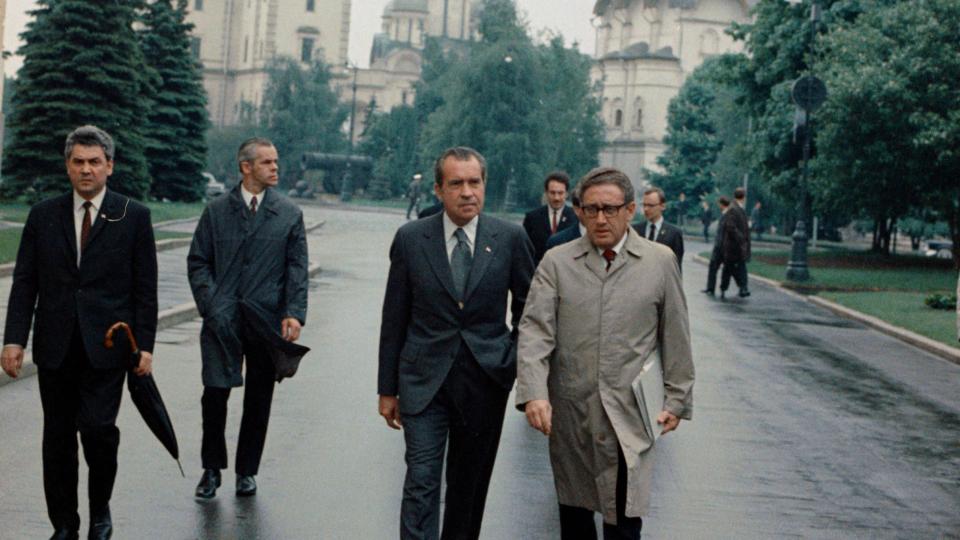 richard nixon and henry kissinger walk on wet pavement near trees and stone buildings, other men walk in the background