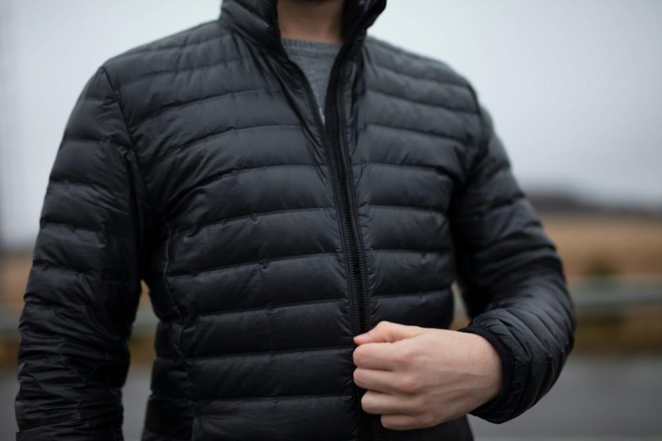 A person shown only from the neck down unzips a black fitted puffer jacket.