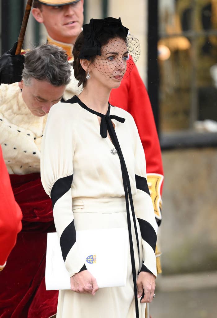 Rose Hanbury in a light dress with trim, a hat with veil, and a clutch at a formal event