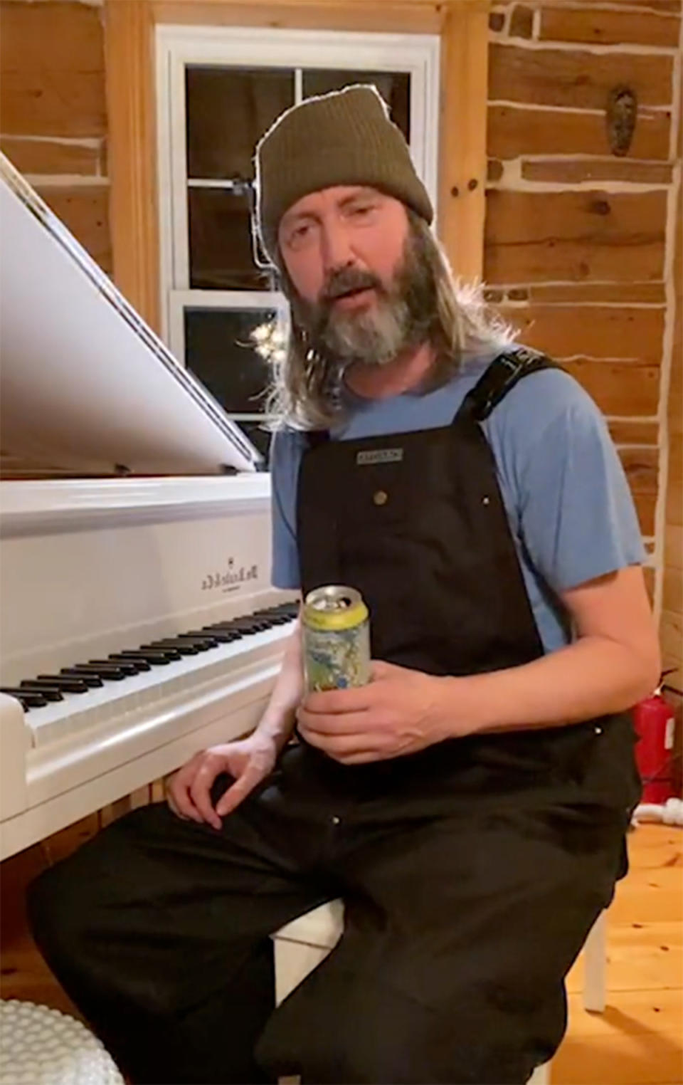 Tom Green wearing black overalls and a green beanie while playing a white grand piano. Photo: Instagram/tomgreen.