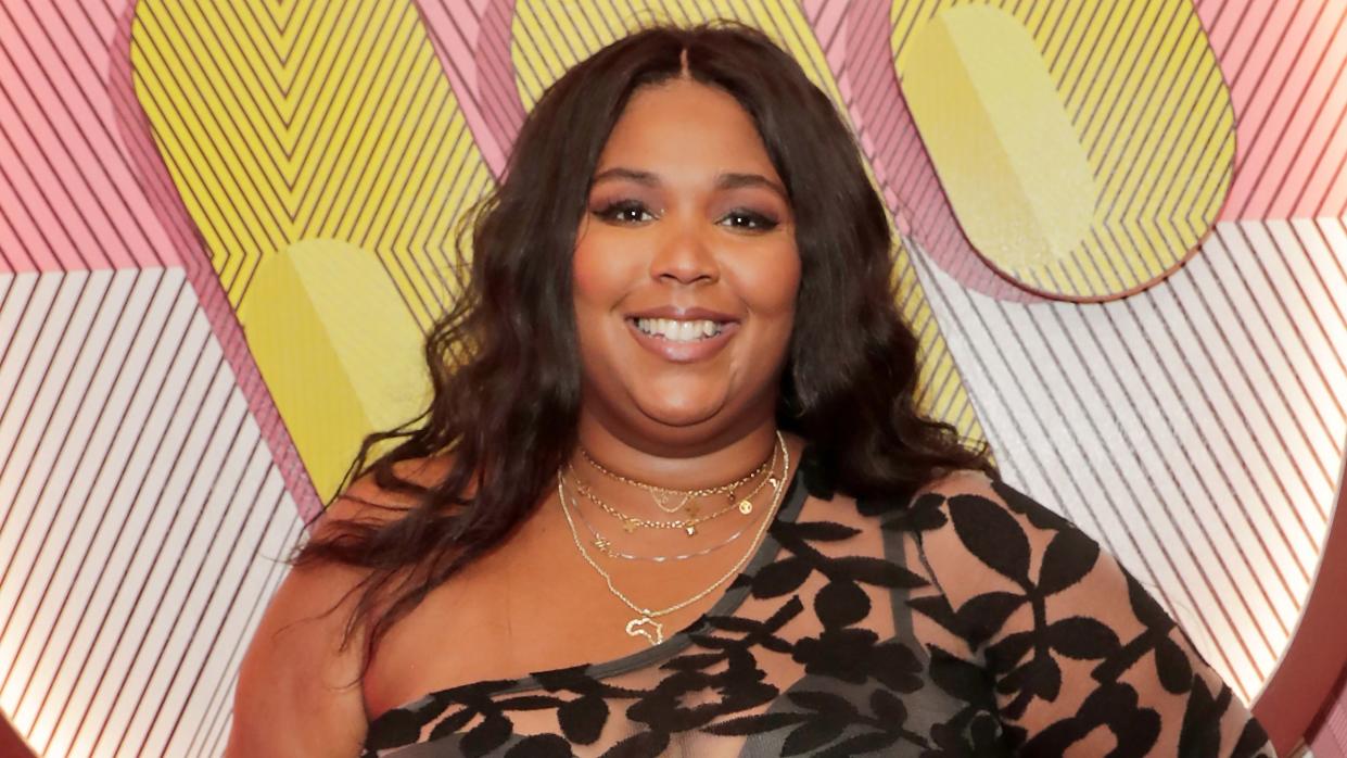 lizzo smiles at the camera, she wears a sheer black floral dress and gold necklaces