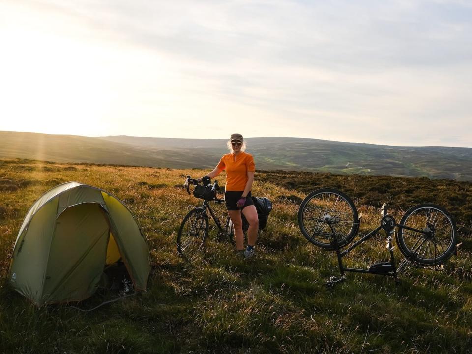 Sarah Morgan near a tent and two bikes with the sun shining behind her.