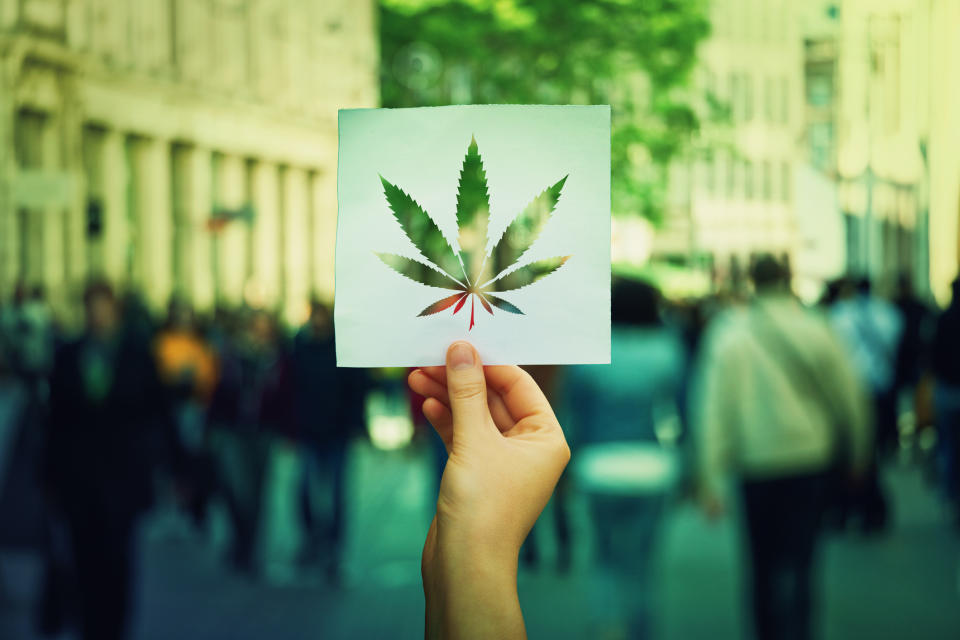 A hand holding up paper sheet with a marijuana leaf cut out of it and a crowded street in the background.