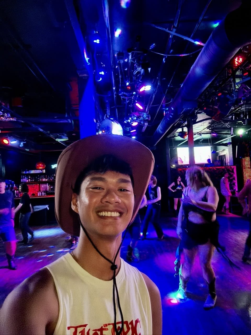 A man wearing a cowboy hat and sleeveless shirt smiles for a selfie at a lively dance club with people dancing in the background