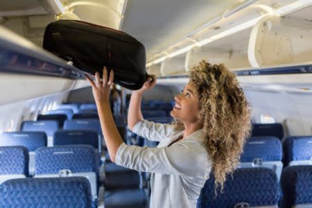 A smiling young businesswoman stands in the aisle of a commercial airliner and places her carry on bag in the overhead bin.