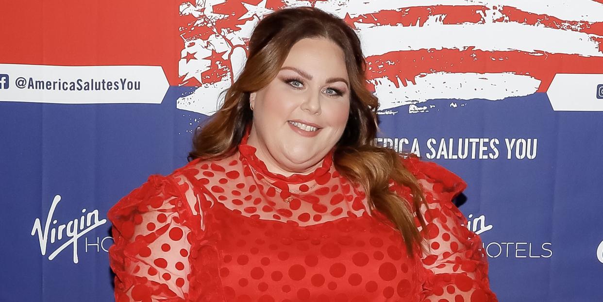 chrissy metz, a woman stands looking and smiling at the camera, she has long brown hair worn in loose waves and wears a red dress with red heels