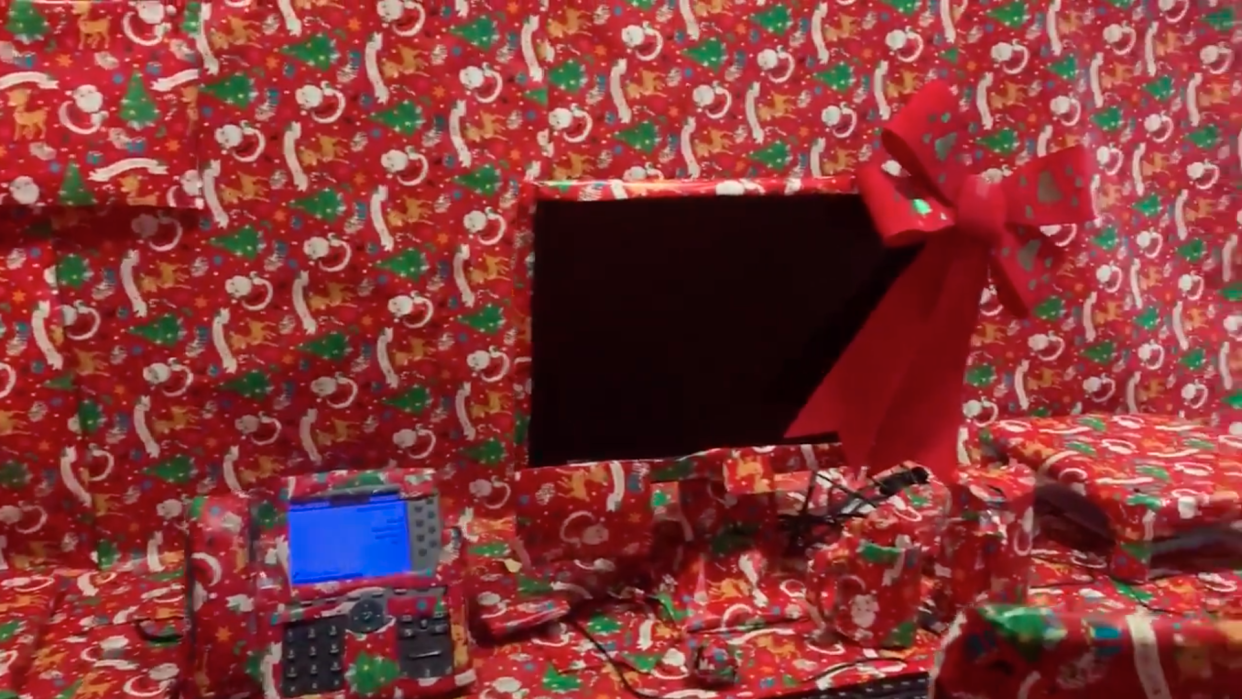 Screenshot of office desk including landline telephone, desktop computer, keyboard and unidentified office supplies wrapped in red christmas wrapping paper, with a red bow.