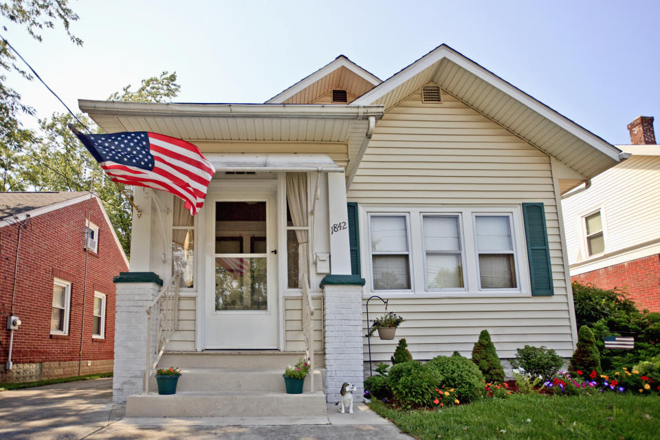 A charming house with an American flag on the porch. A small dog is sitting near the steps. The home has a well-kept lawn with flowers and shrubs