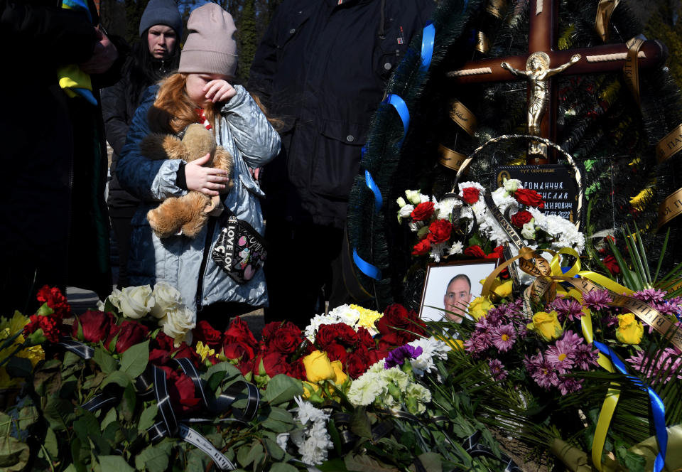 A child and her mother stand near bouquets of flowers and a picture of a soldier who was killed in a Russian airstrike.