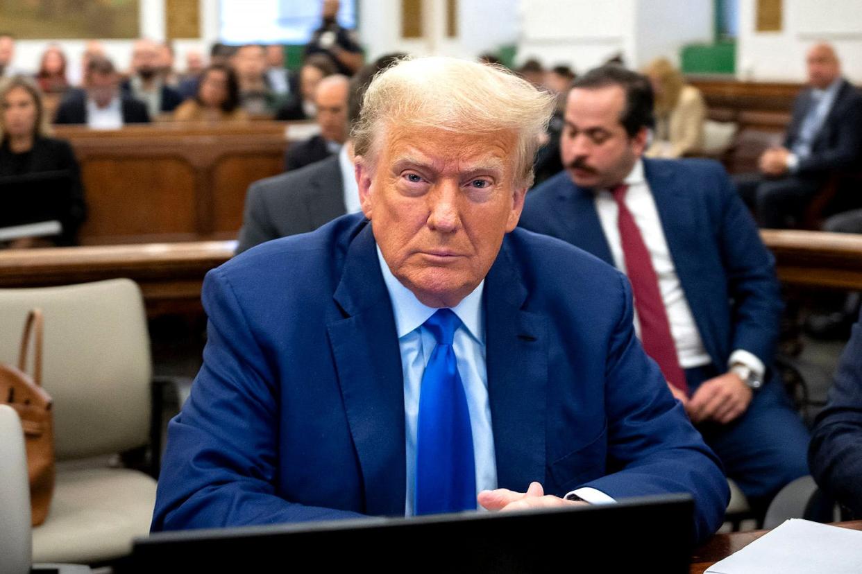 Trump wears a blue suit, blue tie, and blue shirt. He stares straight ahead at the camera, looking almost numb.