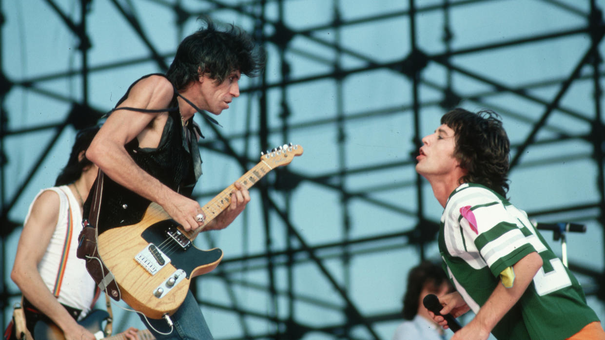  Mick Jagger, wearing a football jersey, interacts with Keith Richards during a Rolling Stones concert. Ron Wood plays guitar beyond. 