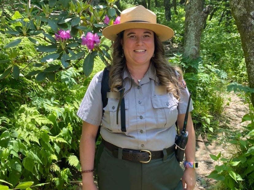 Danielle, wearing a park ranger uniform and carrying a reusable water bottle, walks on a trail between trees and flowers.