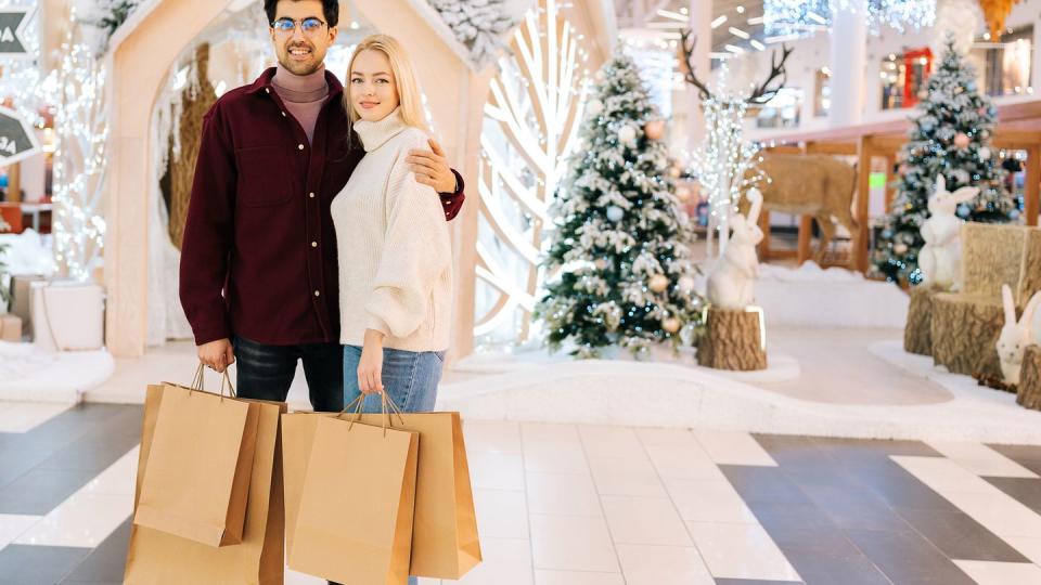 portrait of happy young couple holding shopping paper bags with gifts and purchase looking at camera standing hugging in hall of celebrate shopping mall, with bright xmas decorations in christmas eve