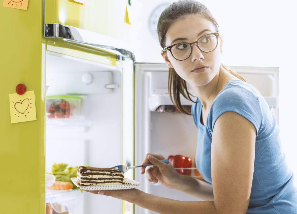 Hungry young woman taking a delicious dessert from the fridge and having a snack, diet fail concept