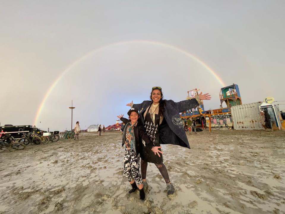 Snyder and her son standing on muddy ground in front of a rainbow at Burning Man 2023.