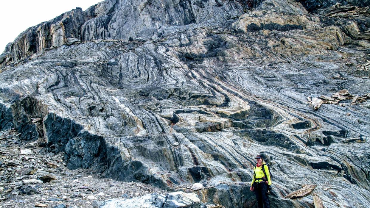  A person with a neon yellow jacket stands in front of a huge wall of rock with cool striations. 