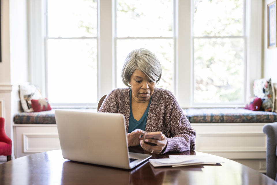 Woman using a laptop and smartphone in a well-lit room with windows in the background