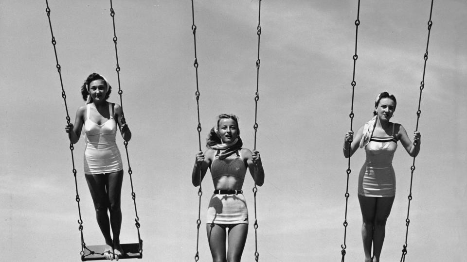 Three women wearing bathing suits and scarves in their hair fly through the air standing up on metal swings, early 1950s