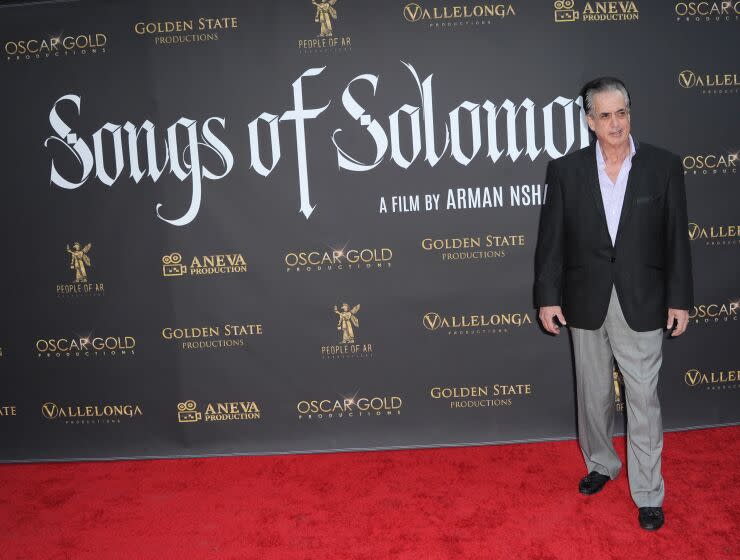 Frank Vallelonga Jr. stands on a red carpet in a black suit and gray dress pants