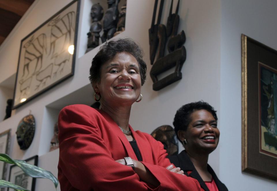dr marilyn gaston smiles down at the camera while standing with her arms crossed, she wears a red suit jacket, gold hoop earrings, a gold necklace, and a gold bracelet, her short hair is styled with loose curls, and her makeup includes soft red lipstick, in the background on the right is dr gayle porter who also has short hair and is smiling while looking away from the camera