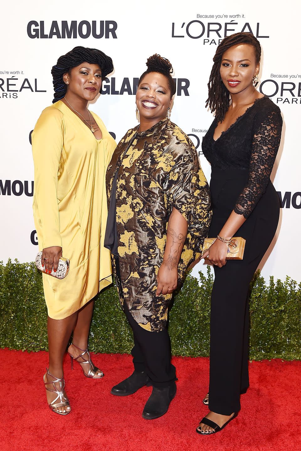 alicia garza, patrisse cullors, and ayo tometi stand on a red carpet smiling and posing for photos, behind is a green shrub and white background with logos, garza wears a yellow knee length dress, cullors wears a yellow and black jacket with black pants, and tometi wears a black lace shirt and black pants