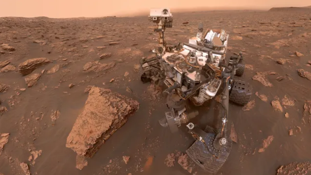  Image of a large whitish rover on the surface of mars, surrounded by rocks and red dirt. 