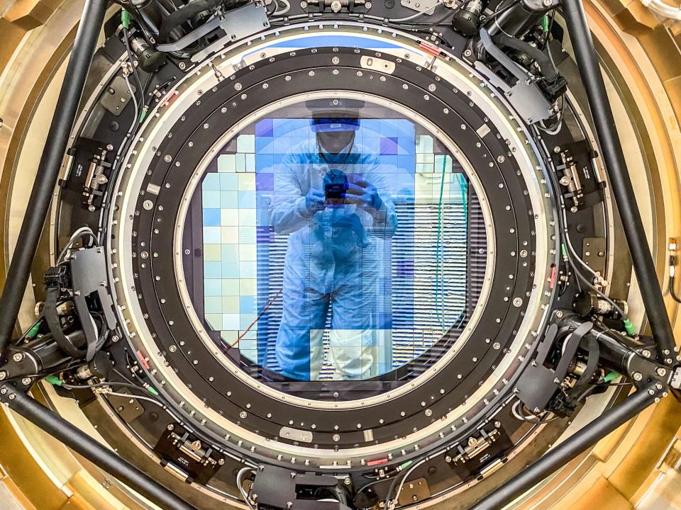 person in white jumpsuit wearing medical gloves appears faint taking selfie in a mosaic of blue green and yellow tiles inside a reflective lens inside a circle of mechanical gear with a gold circular frame around it