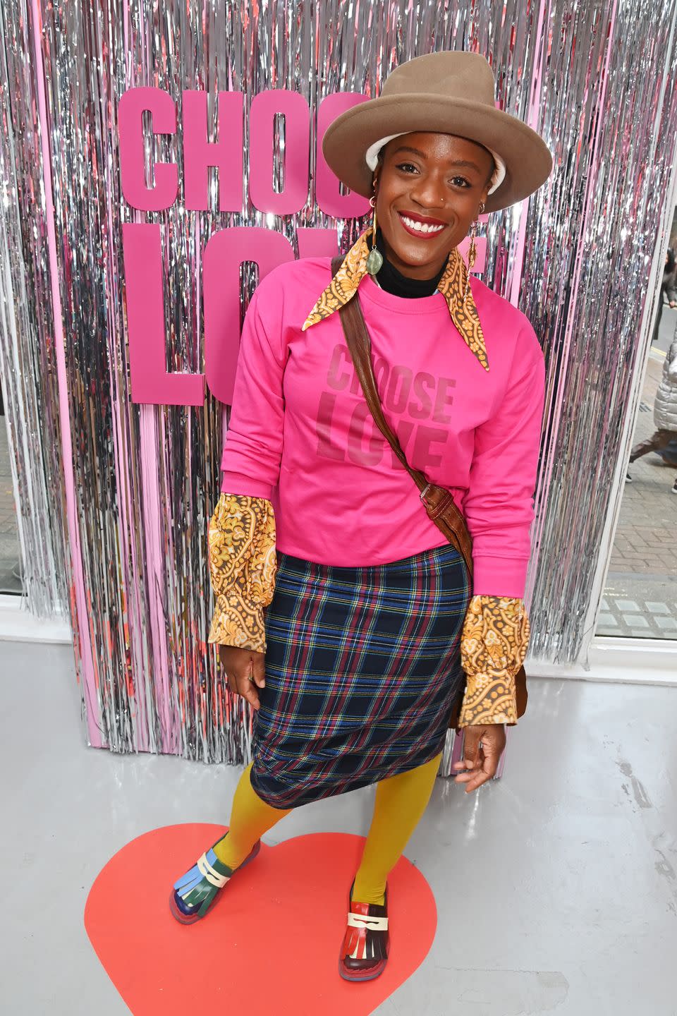 tnia miller, a woman wearing a bright pink sweater and a large fedora hat, standing in front of a wall of silver streamers and text that reads choose love, smiling for the camera