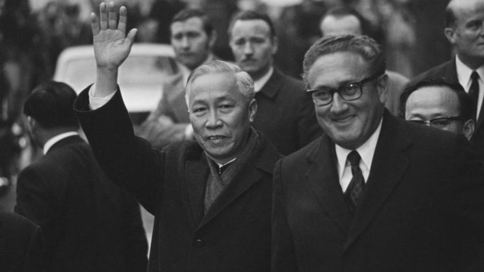 le duc tho waves and smiles while walking on a street next to henry kissinger, also smiling
