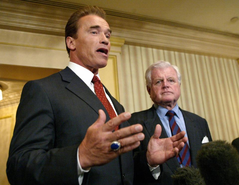 arnold schwarzenegger talking to reporters offscreen in front of microphones with ted kennedy watching behind him