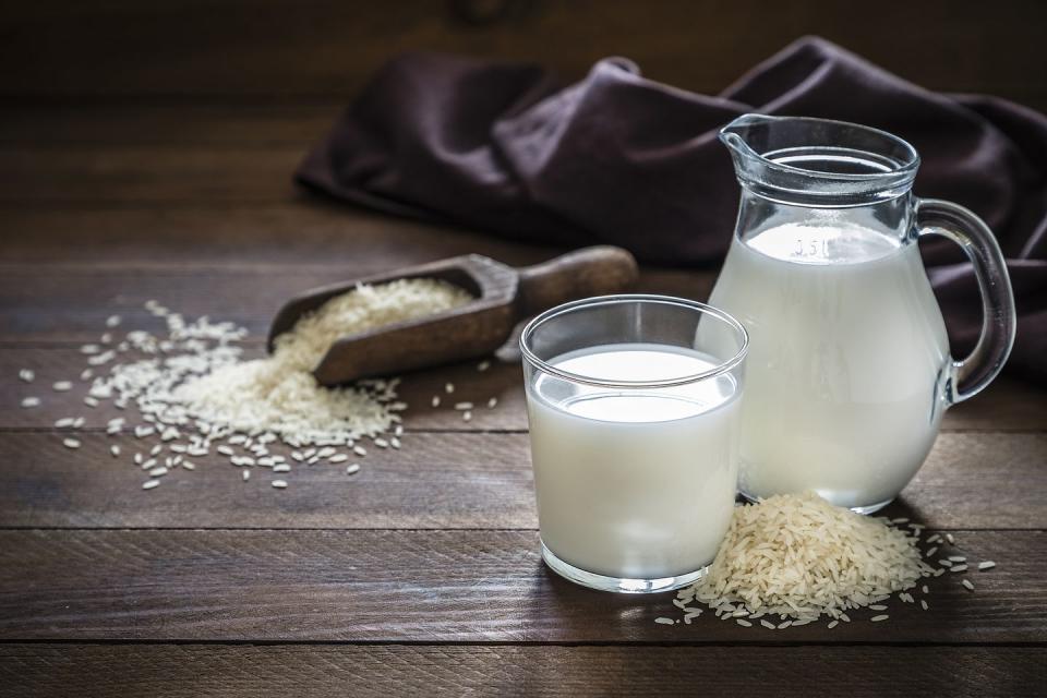 front view of a jug and a drinking glass full of rice milk the glass and the jug are at the right side of the image on a rustic wooden table, next to the glass its a heap of rice grains this image is a part of a vegan milk series low key dslr photo taken with canon eos 6d mark ii and canon ef 24 105 mm f4l