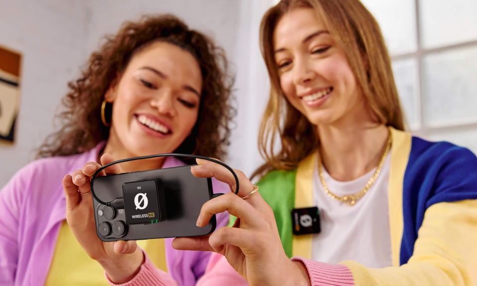 Product lifestyle marketing photo for the Rode Wireless ME mic. Two young people smile as they look at a phone with a receiver attached to its back. One is wearing a wireless mic.