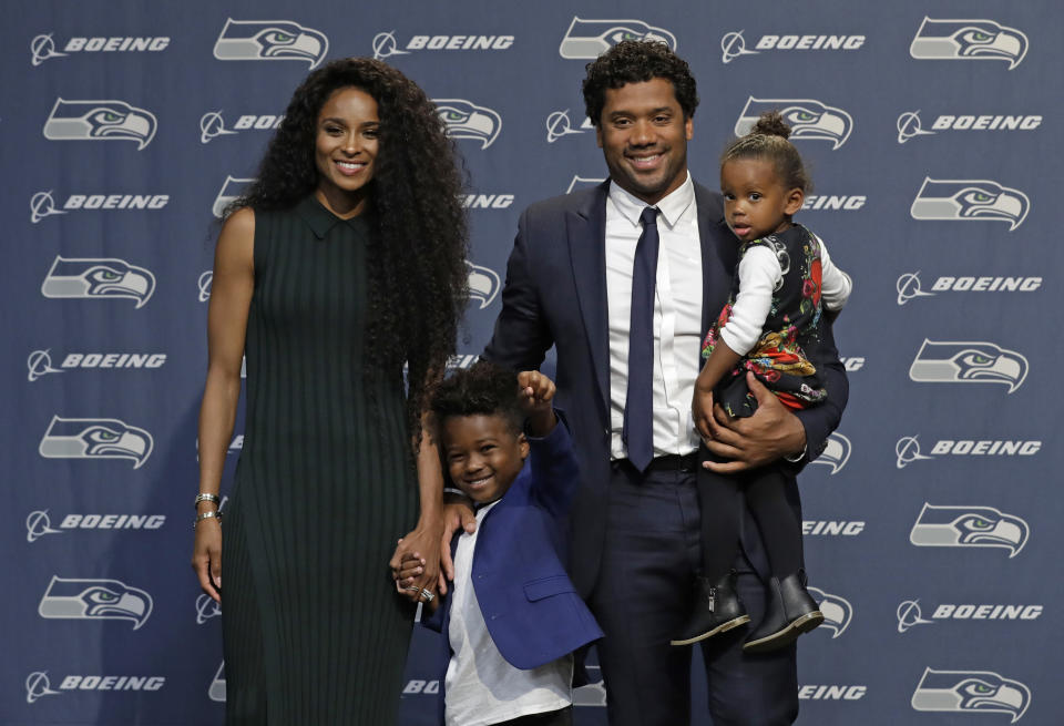 Ciara, Russell Wilson and their two children standing in front of a Seahawks background. 