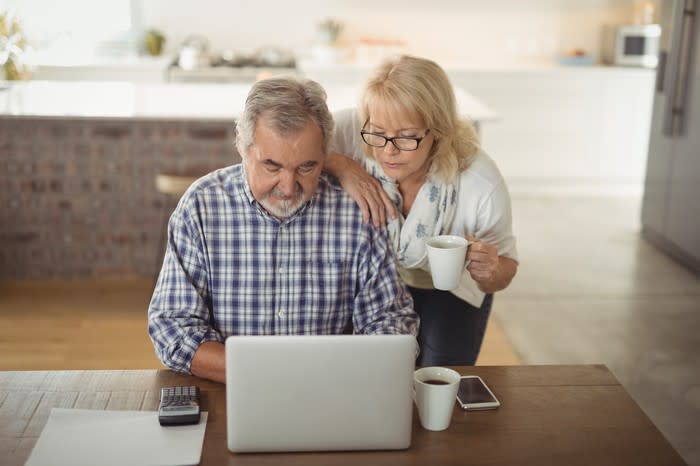 Older woman with mug in hand looking over man's shoulder as he types on laptop