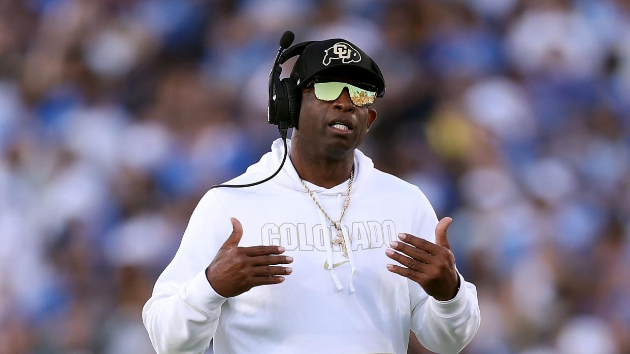 deion sanders gesturing with his hands as he speaks on a headset during a football game