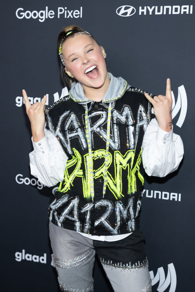 JoJo Siwa poses with a peace sign, wearing a jacket with "KARMA" and sparkly trousers