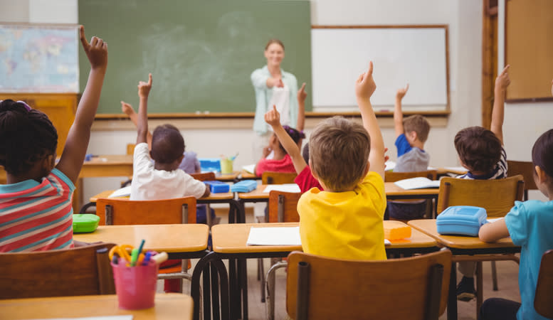 Children raising hands in a classroom with a teacher at the chalkboard