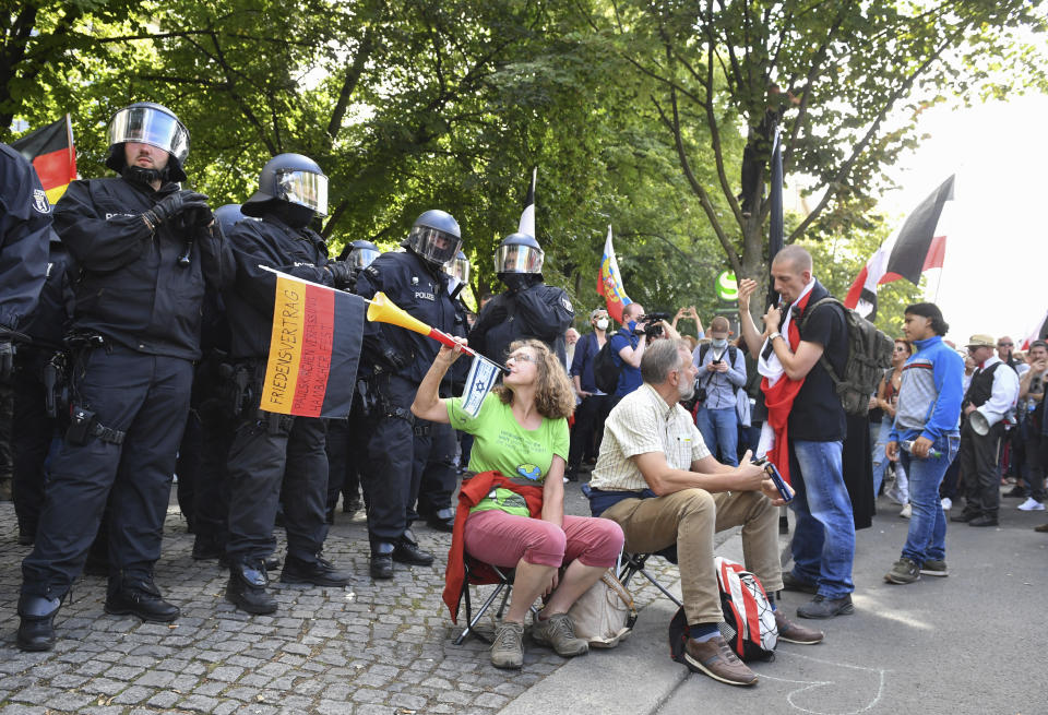 The participant in a demonstration against the Corona measures blows into a vuvuzela with a German and an Israeli flag in front of a police chain in Berlin, Germany, Saturday, Aug. 29, 2020. (Bernd Von Jutrczenka/dpa via AP)